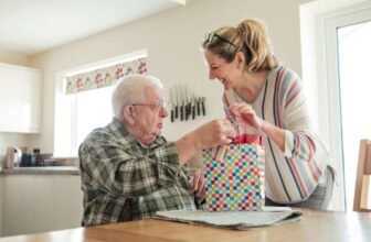 Handing over a gift bag to elderly man during birthday celebration at home, family gift exchange, senior care, happiness, special occasion, love, family bonding, generational connection, celebration, togetherness.