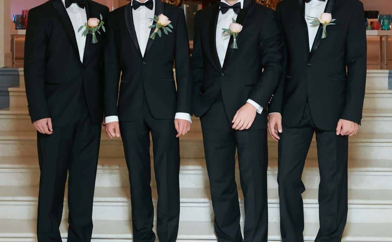 Dapper men in black tuxedos with boutonnieres standing on steps at formal event.