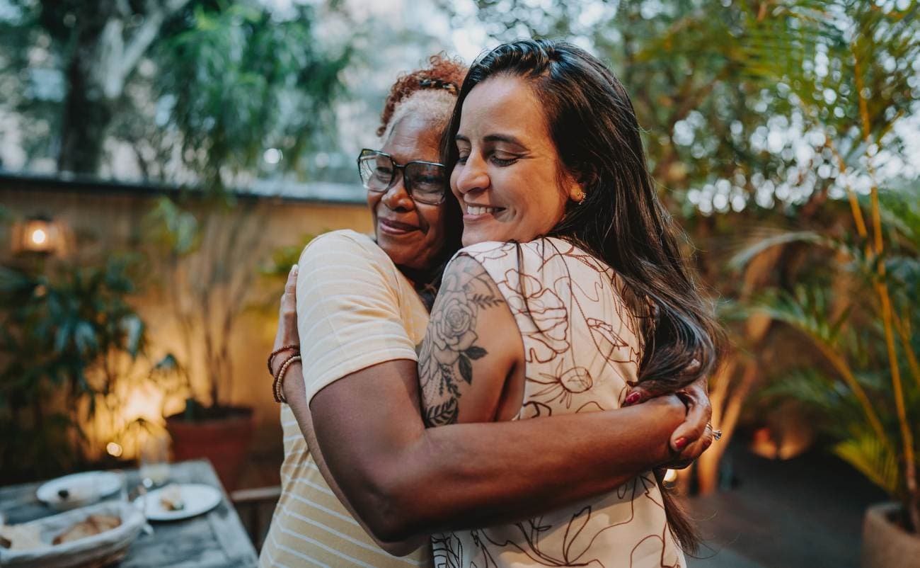 Smiling women hugging outdoors at a garden party, celebrating sale is Live event, women sharing joyful moment, diverse friends enjoying shopping and discounts, happy lifestyle, Sale is Live banner visible in store or online setting.