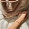 Close-up of a woman's hand wearing a delicate gold bracelet, resting on a soft fabric surface with a beige velvet bag in the background, perfect for Mother's Day gift inspiration.
