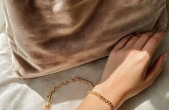 Close-up of a woman's hand wearing a delicate gold bracelet, resting on a soft fabric surface with a beige velvet bag in the background, perfect for Mother's Day gift inspiration.